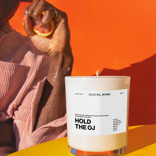 Candle labeled 'Hold the OJ' with a person squeezing a lemon against an orange background